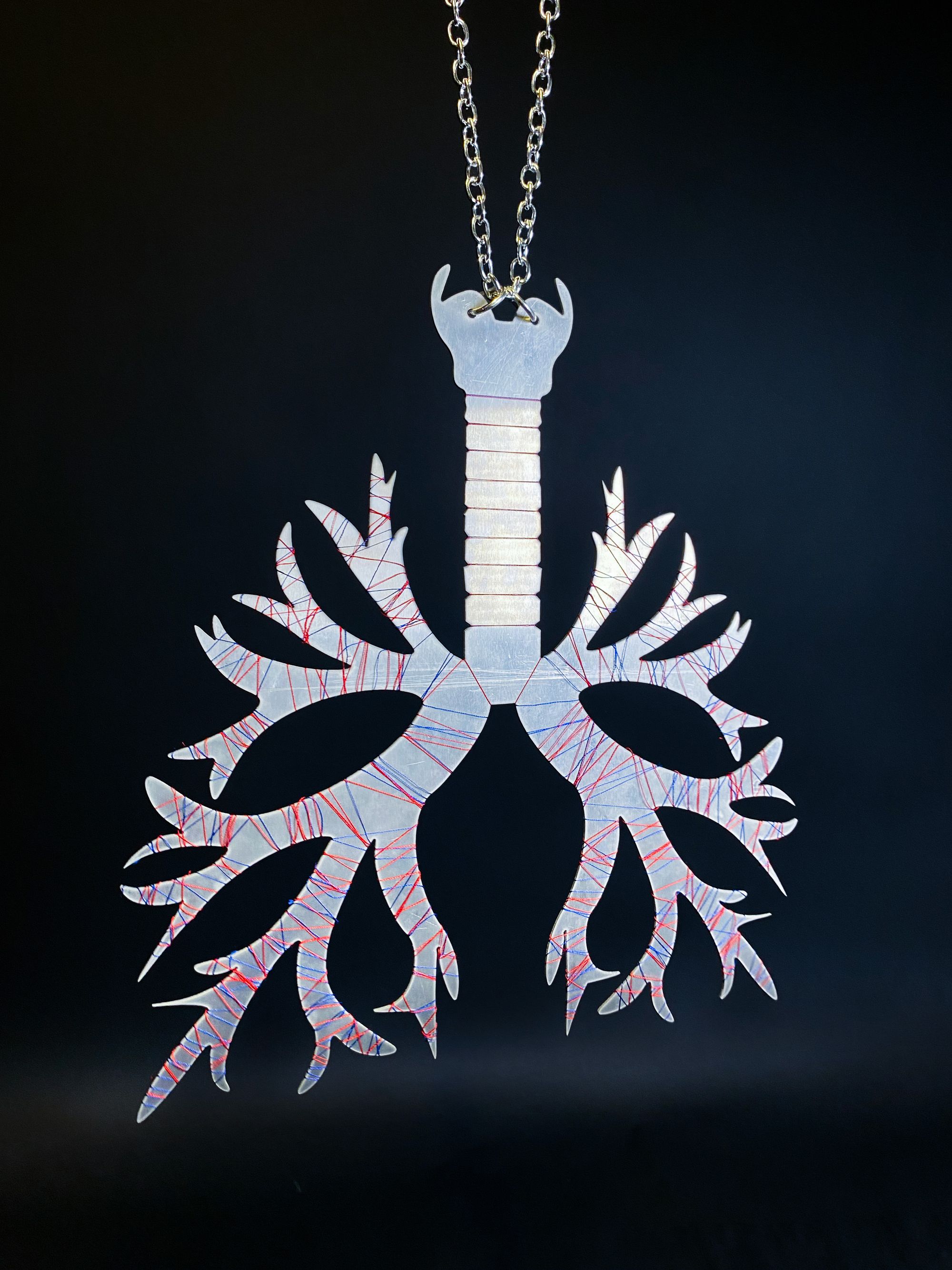 Close-up of a pendant shaped like a pair of lungs with branching bronchi, made from silver-toned metal with red and blue etched lines. The pendant hangs on a chain against a black background.